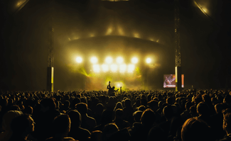 Una gran multitud de personas se enfrenta a un escenario brillantemente iluminado en un concierto bajo techo, con luces amarillas que brillan a través del humo y una persona elevada por encima de los hombros de la multitud en el centro.