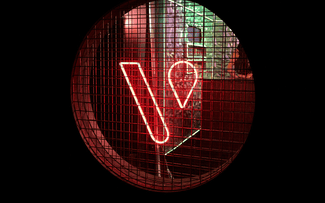 A través de una rejilla metálica circular se ve un letrero de neón rojo con la letra V, con un interior poco iluminado y luces verdes y rojas de fondo.