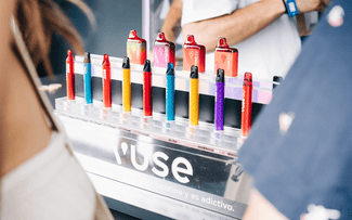 Un expositor que muestra coloridos bolígrafos para vapear Vuse en fila, cada uno con el cuerpo de un color diferente, con personas difuminadas en primer y segundo plano. El expositor incluye una etiqueta de advertencia en español.